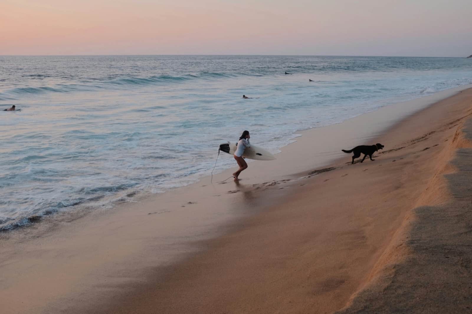 A surfer heading into the waves with a dog running beside her on the beach at San Pancho, Nayarit
