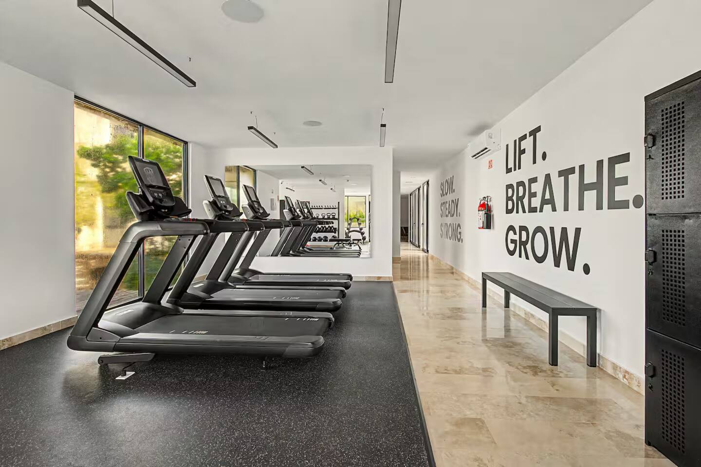 El gimnasio cuenta con varias caminadoras para cardio, además de mancuernas, pesas libres, bancos de pesas, equipo de fuerza y más, todo en un espacio luminoso con aire acondicionado, grandes ventanales y luz natural.