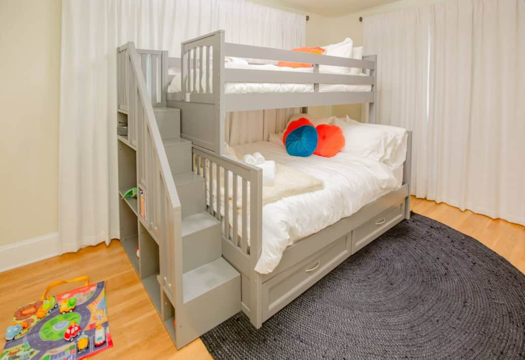 A cozy full-size bottom bunk topped with a twin upper, both featuring extra-comfortable mattresses. At the end of the bed, built-in shelves hold a selection of toys and books perfect for little ones ages 18 months to 5 years.