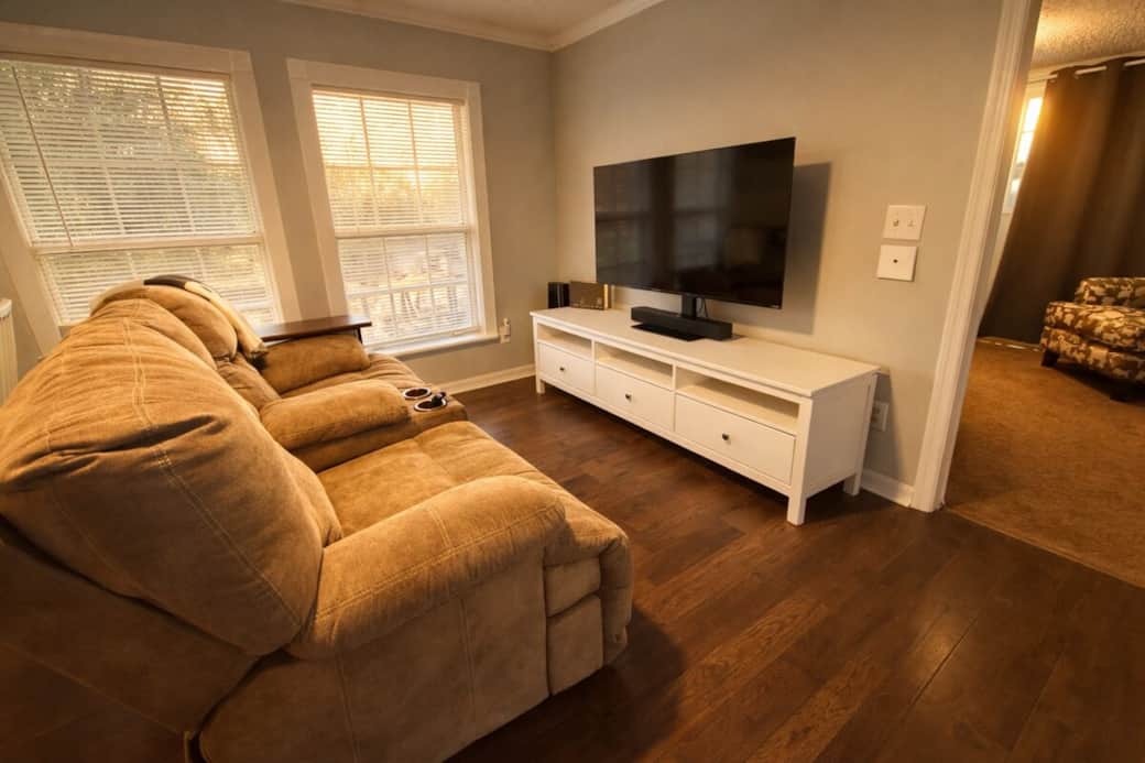 Kick back in a bright, cozy living room featuring a plush reclining sofa and a large flat-screen TV. Warm wood floors and soft natural light from two big windows make it an easy spot to relax, stream your favorites, or unwind after a day out.