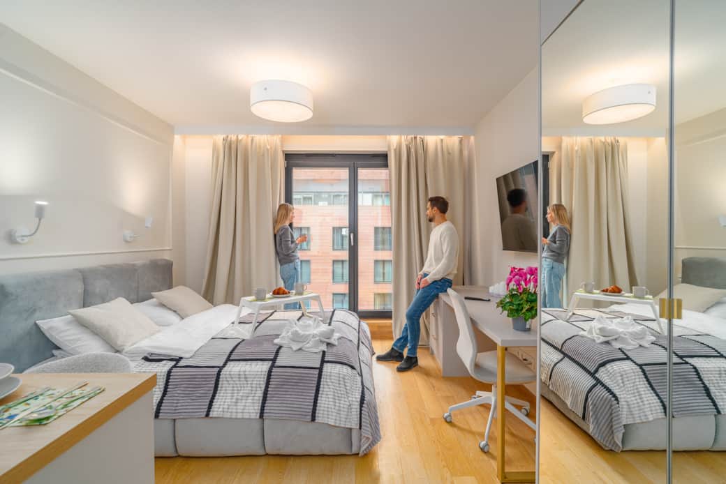 A modern, bright hotel room with a large bed and a couple talking by the window.
