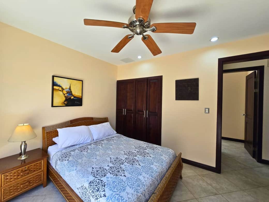 Comfortable guest bedroom with queen bed, ceiling fan, and warm natural tones. Features built-in closet, dresser, and easy hallway access for added convenience.
