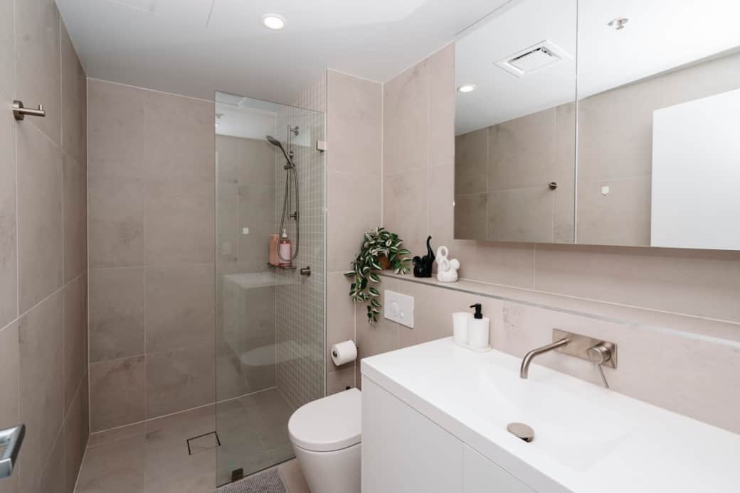 This contemporary bathroom is designed with your comfort in mind. Featuring a pristine white palette, it includes a walk-in shower, a sleek sink, and a modern toilet. The large mirror and well-placed lighting enhance the sense of space.