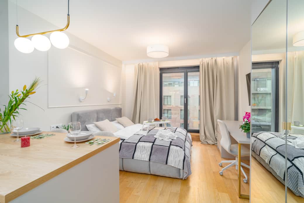 A bright studio apartment with a kitchenette, a large bed, and a balcony window.
