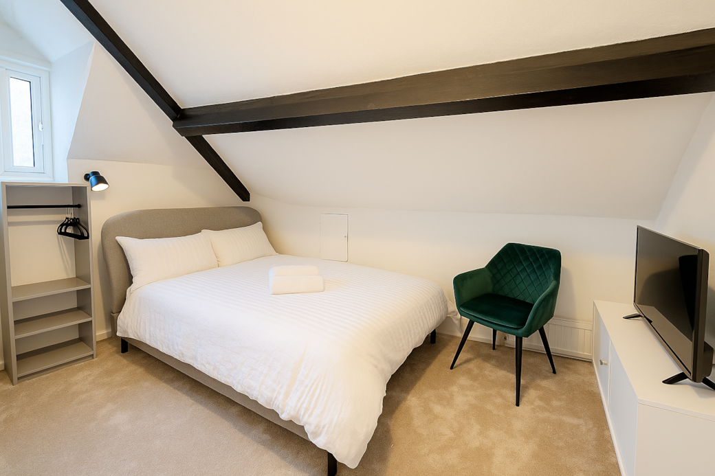 Charming attic bedroom with exposed beam and plush bedding, complete with TV, velvet green armchair and open wardrobe. The sloped ceiling and soft lighting create a warm, inviting atmosphere.