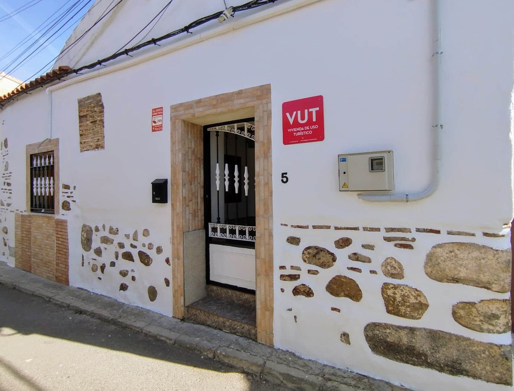 Fachada de vivienda de uso turístico con paredes blancas, detalles en piedra y ladrillo, puerta con reja metálica y cartel VUT visible