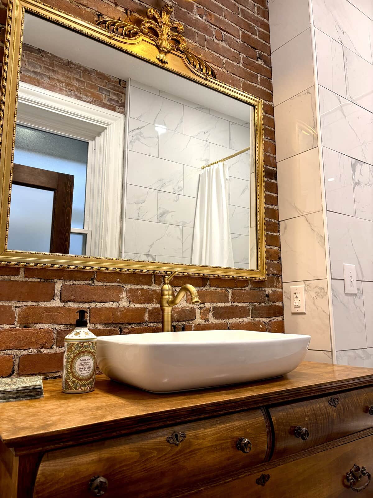 A vessel sink and brass faucet on an antique dresser, set against original exposed brick