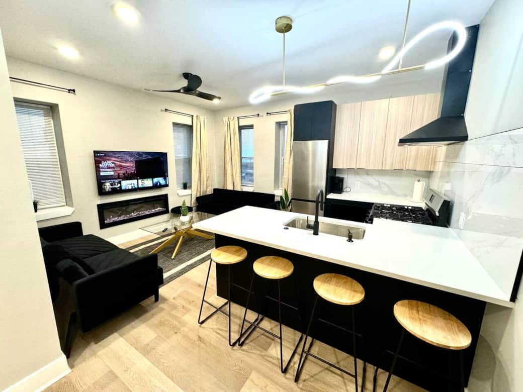 Modern kitchen featuring brand-new appliances, including a breakfast bar, dishwasher, microwave, and refrigerator. It seamlessly connects to the living room, which is furnished with 2 sofa beds, fireplace ,and a flat-screen TV.
