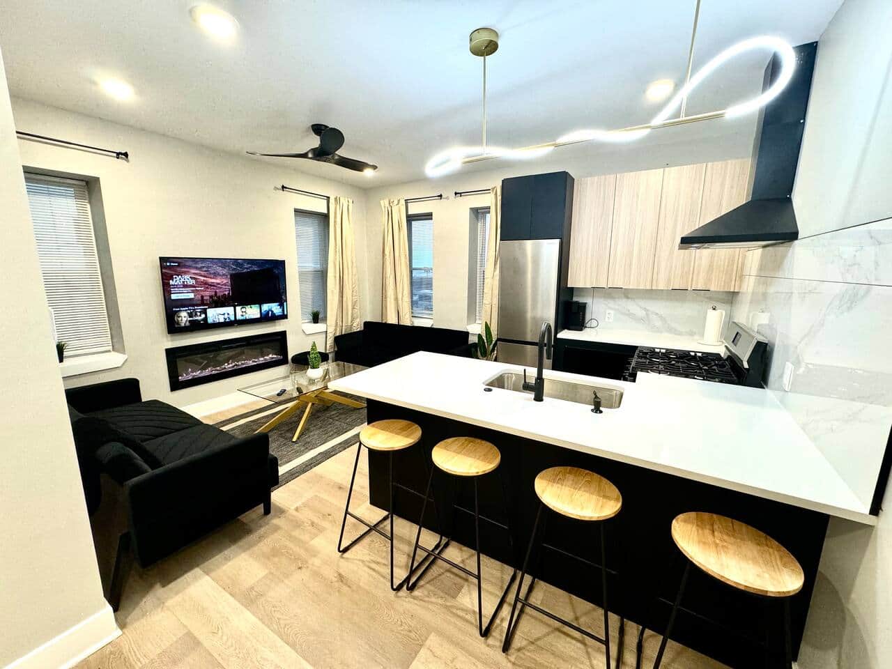 Modern kitchen featuring brand-new appliances, including a breakfast bar, dishwasher, microwave, and refrigerator. It seamlessly connects to the living room, which is furnished with 2 sofa beds, fireplace ,and a flat-screen TV.