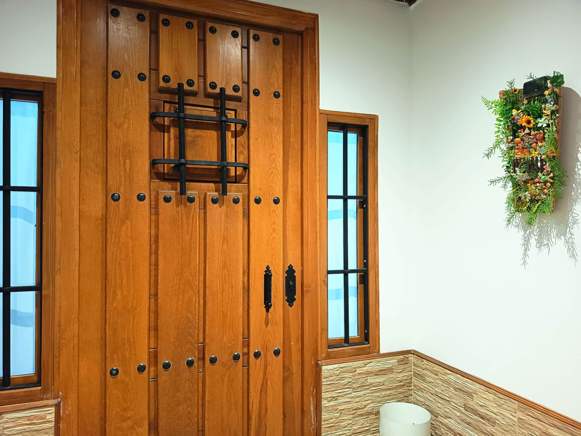 Puerta de madera rústica con herrajes negros, flanqueada por ventanas con rejas y decoración floral en la pared