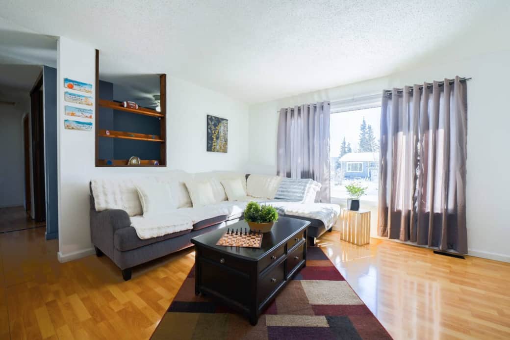 A bright living room with large windows, a cozy sectional sofa with soft throws, and a stylish coffee table with a chess set. Enjoy the natural light, wooden floors, and a peaceful space to relax.