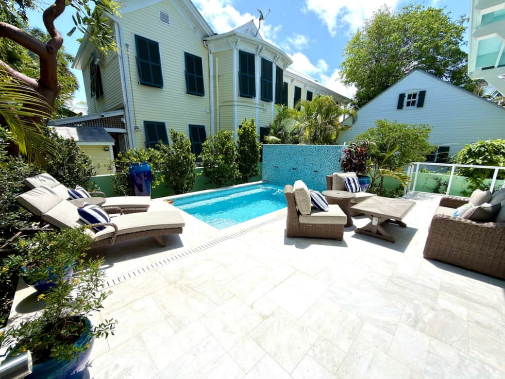 A private outdoor retreat centered around a saltwater pool containing a waterfall feature surrounded by an outdoor shower, lounge chairs, tables, sofa, 2 sitting chairs, and a TV. Designed for relaxing, gathering, and enjoying the Key West sunshine.