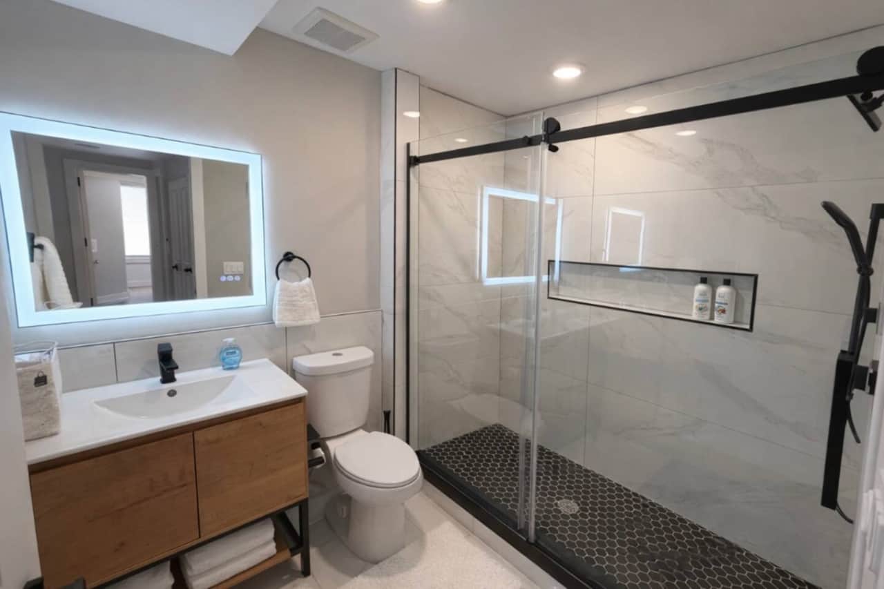 Modern bathroom featuring a beautifully tiled Carrara shower, glass enclosure, and a sleek vanity with a backlit mirror for a bright, spa-like feel.