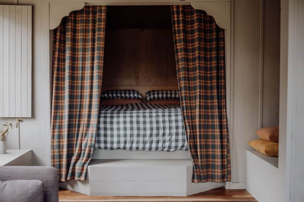 A serene bed nook featuring a plush king-sized bed draped in soft, luxurious linens. A step assists in getting in and out of the bed, with block out flannel curtains & benches on either side.