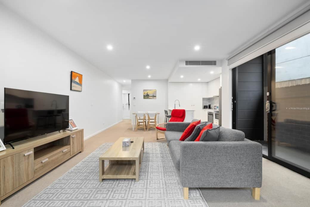 Step into the living area ,the gray sofa and red chair offer a pop of color, while the wooden coffee table and TV stand add a touch of elegance. Large windows provide ample light and a glimpse of the serene backyard, creating a peaceful atmosphere.