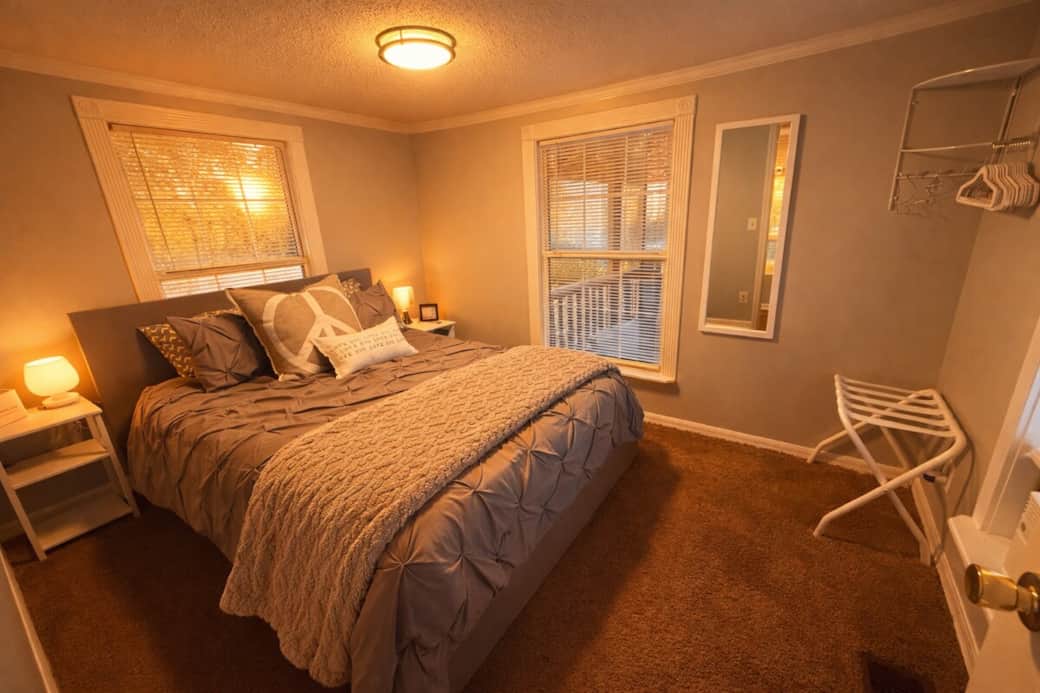 Unwind in a calm, comfortable bedroom with a cozy bed, soft linens, and warm bedside lighting. Large windows bring in natural light, and a window A/C unit keeps things cool and comfortable for sleeping.