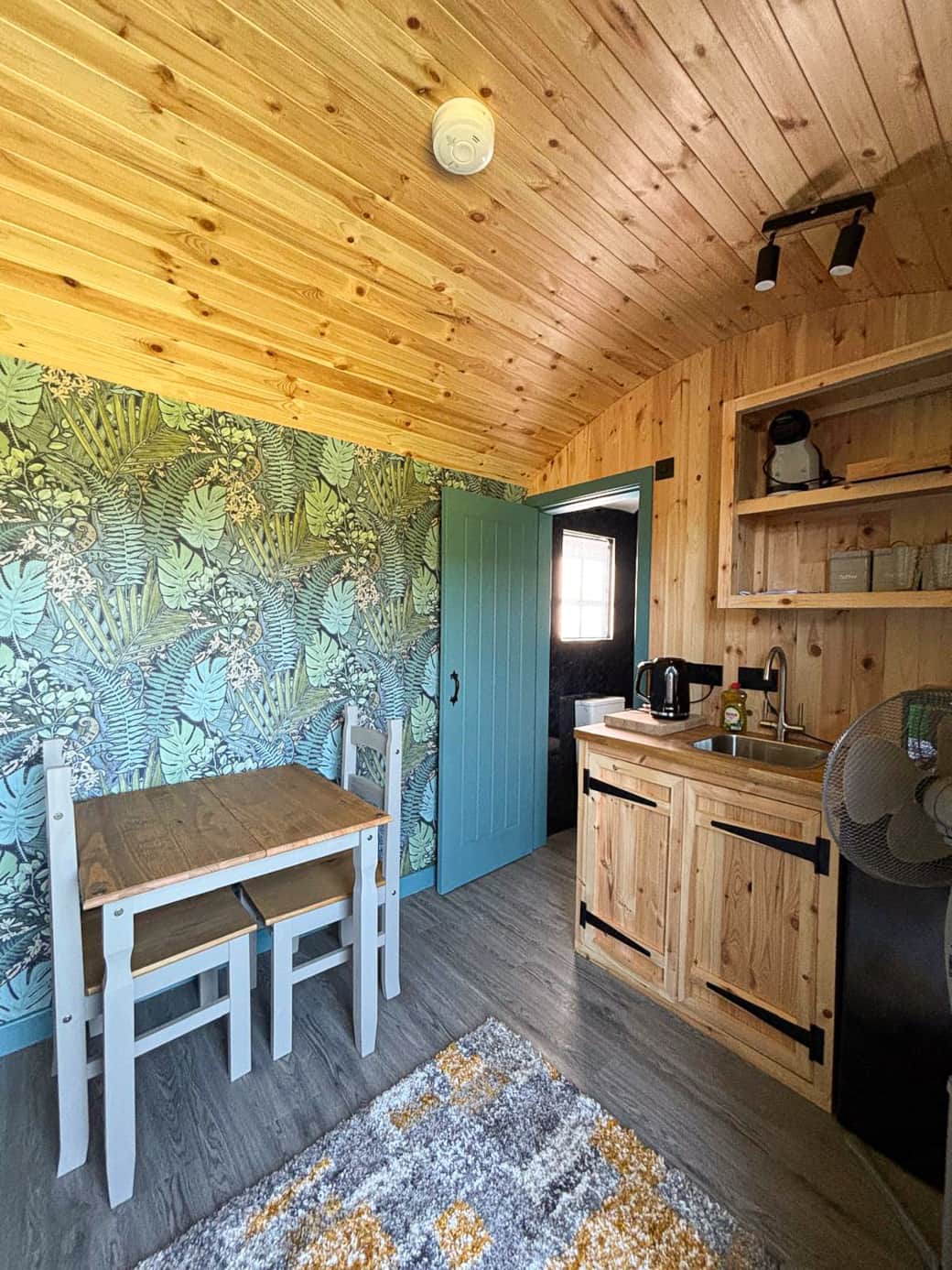 Compact dining and kitchen space with natural wood finishes and leafy feature wall.