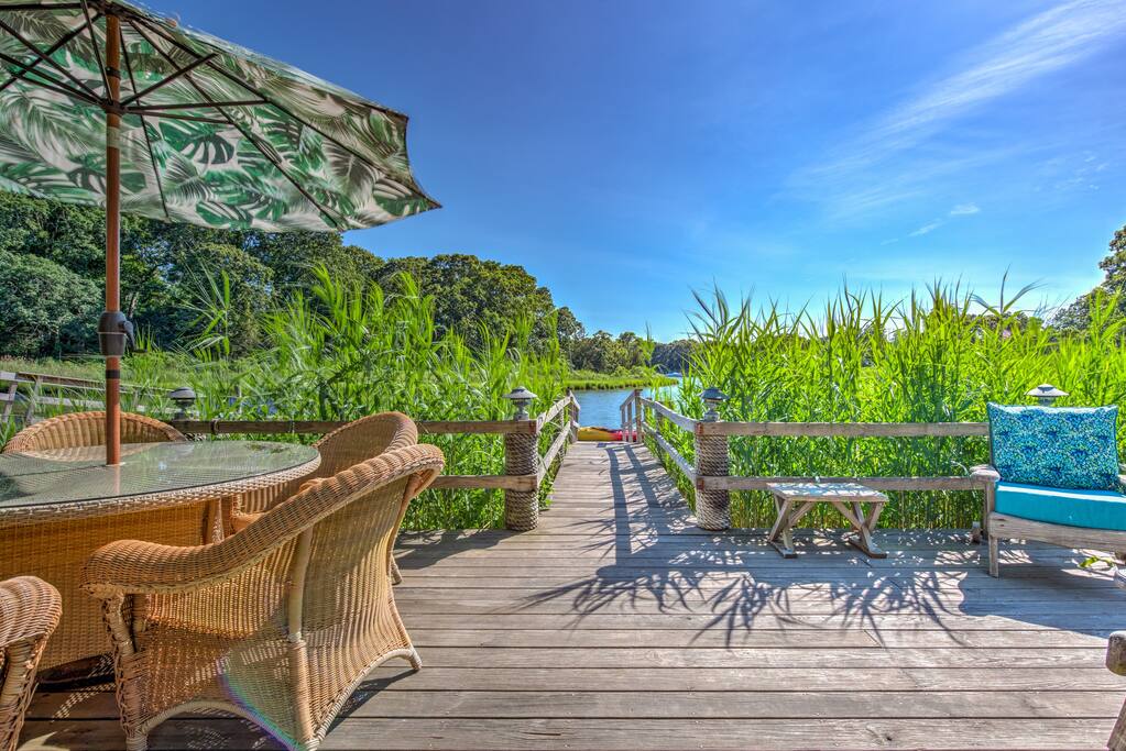 Dining table under an umbrella on the dock, with beach grass and the cove beyond