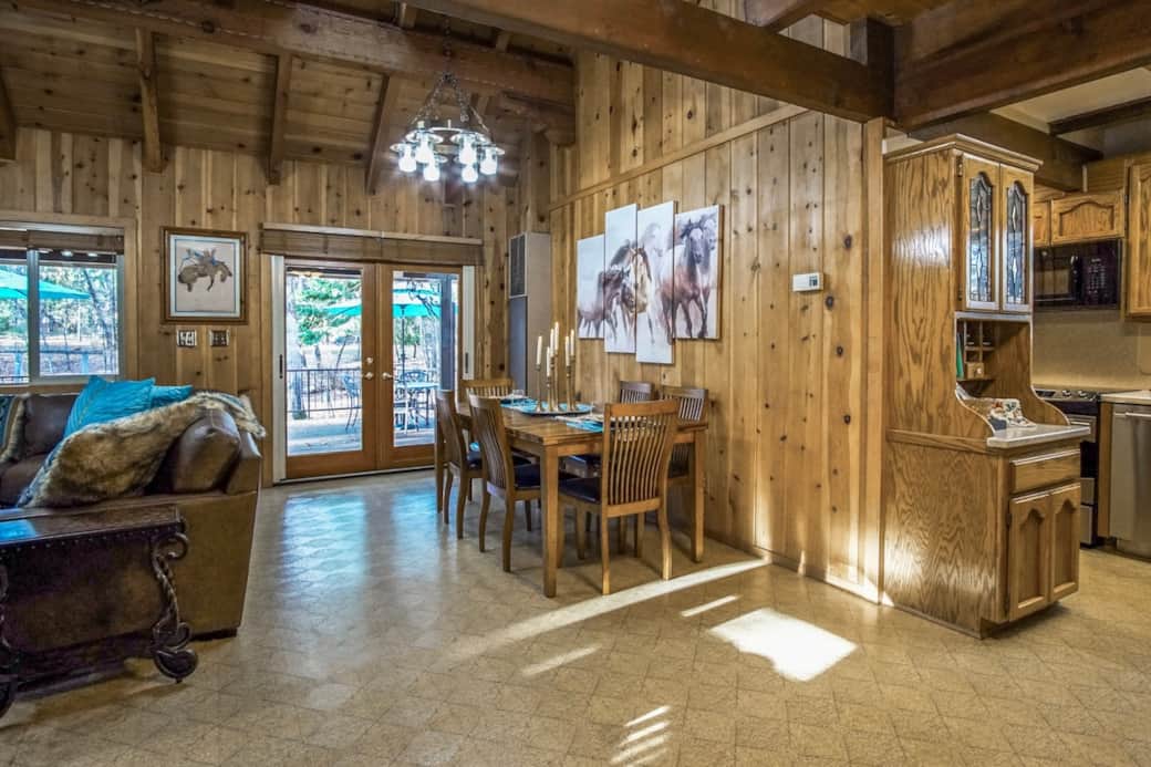 Bright, open great room with warm wood walls, horse art, rustic chandeliers, and bold turquoise accents—flowing from dining to living and out to the deck through French doors.