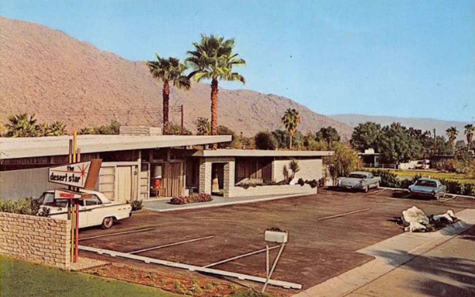A mid-century modern hotel building with a flat roof, a parking lot, and palm trees in front of a mountain view.