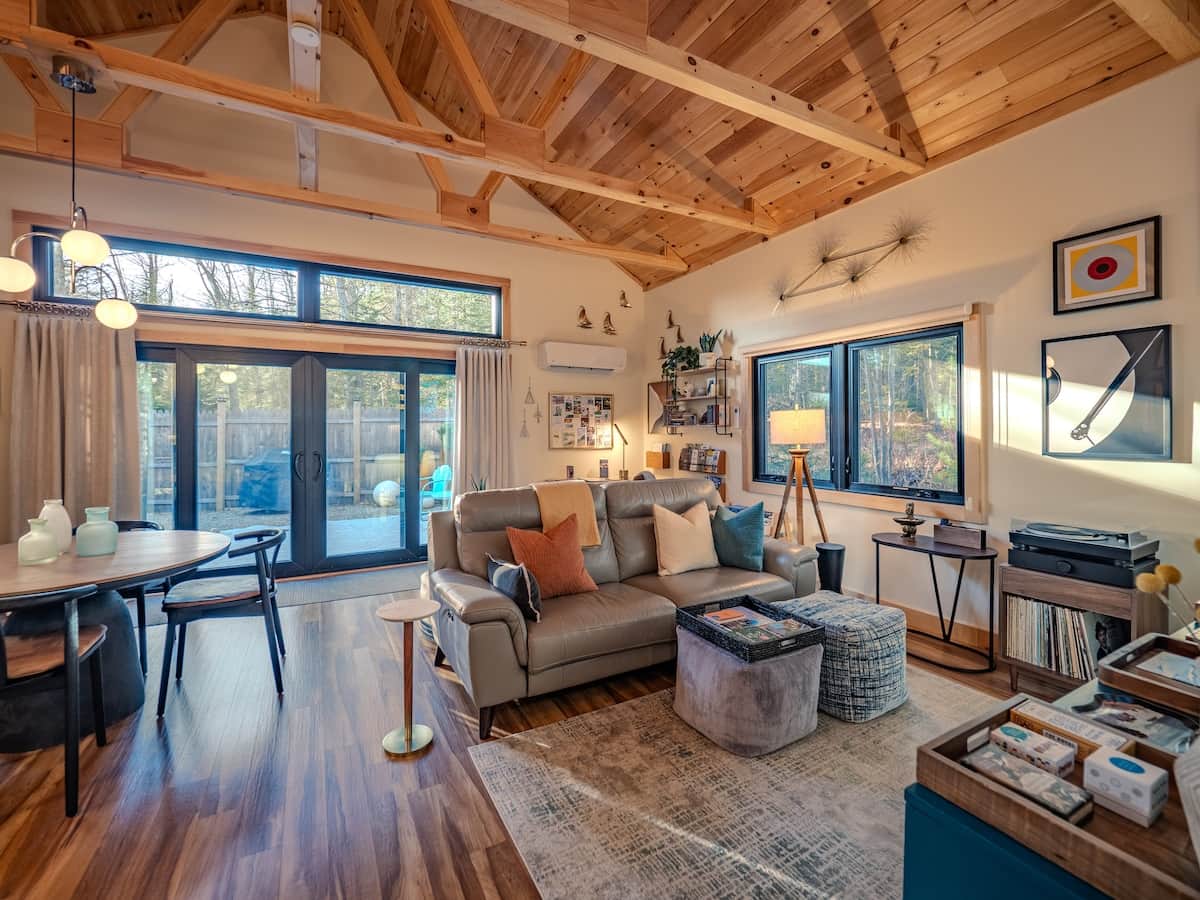 This warm, inviting open-concept living area blends rustic cabin architecture with mid-century modern styling. The space is defined by a dramatic vaulted ceiling with exposed pine beams and large windows that flood the room with natural light.