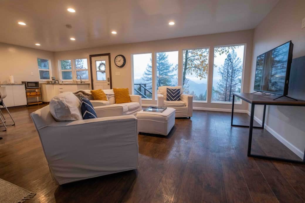 Living room with kitchenette behind it. Notice ALL the windows. Look out at the trees and mountains. We have added white floor to ceiling curtains on the large windows. 
