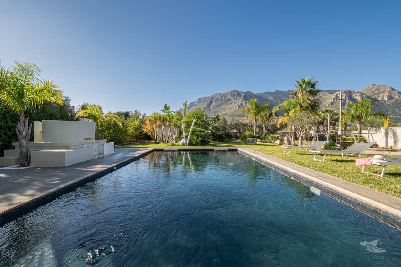 The villa’s pool sparkles under the clear sky, framed by vibrant palm trees and majestic mountains.