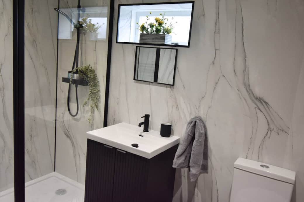 A sleek, modern bathroom featuring marble-effect walls, a walk-in rainfall shower, matte black fixtures, and fresh greenery. The space is bright, stylish and ideal for a relaxing start or end to your day.