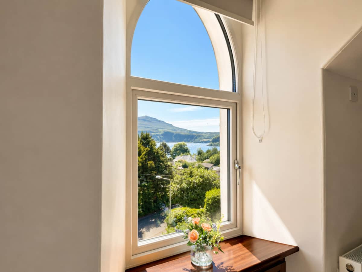 Arched upstairs window framing treetops, Loch Portree and the hills beyond.