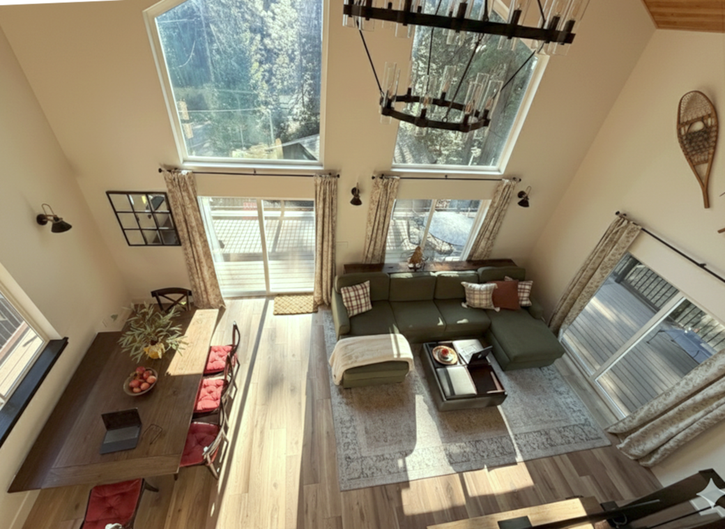 Spacious, light-filled living room with vaulted ceilings and large windows showcasing forest views. Features a comfy sectional, dining area,  Smart TV over the fireplace, plus easy deck access—perfect for relaxing, movie nights, or family gatherings.