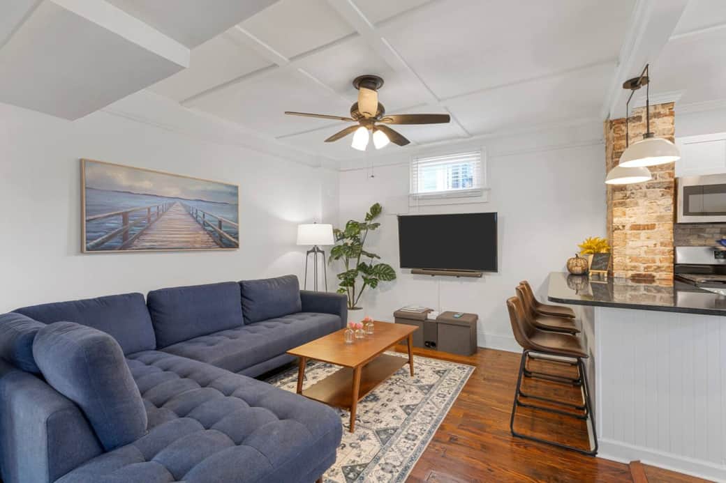 Living room with a large inviting cloth couch, Sony 55" HDTV/smart TV with built-in streaming ability and Polk Audio Sound bar with Sub. Bar chairs, Floor lamp, and extra blankets stored in the grey floor cushioned box seats.  3 bar stools at counter