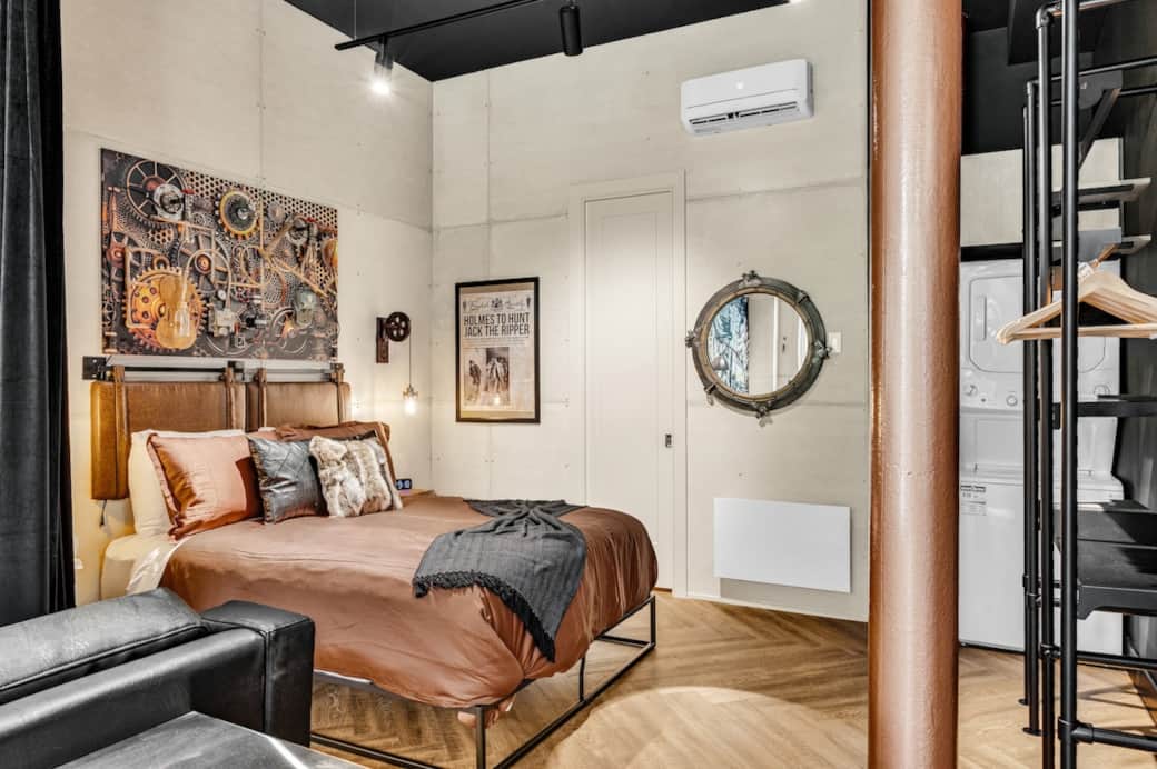 Espace chambre stylisé ; Séparée de l’aire commune par un rideau de velours noir, l’espace allie intimité et élégance théâtrale.
Lit ultra-confortable, sous une œuvre murale steampunk fait par un artiste, une penderie ouverte façon boutique.
