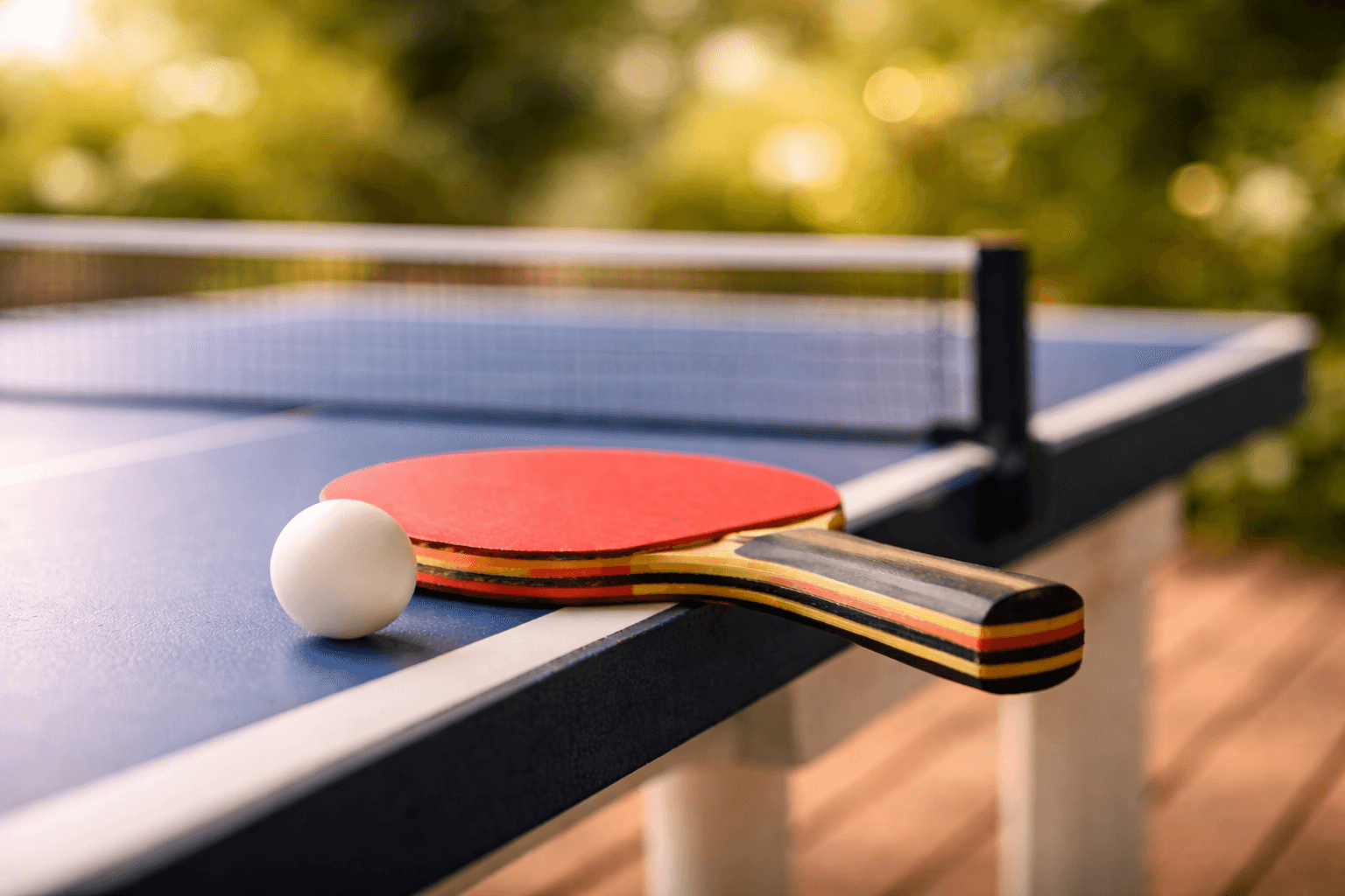 Table tennis bat and ball on an outdoor table tennis table, with blurred greenery in the background.