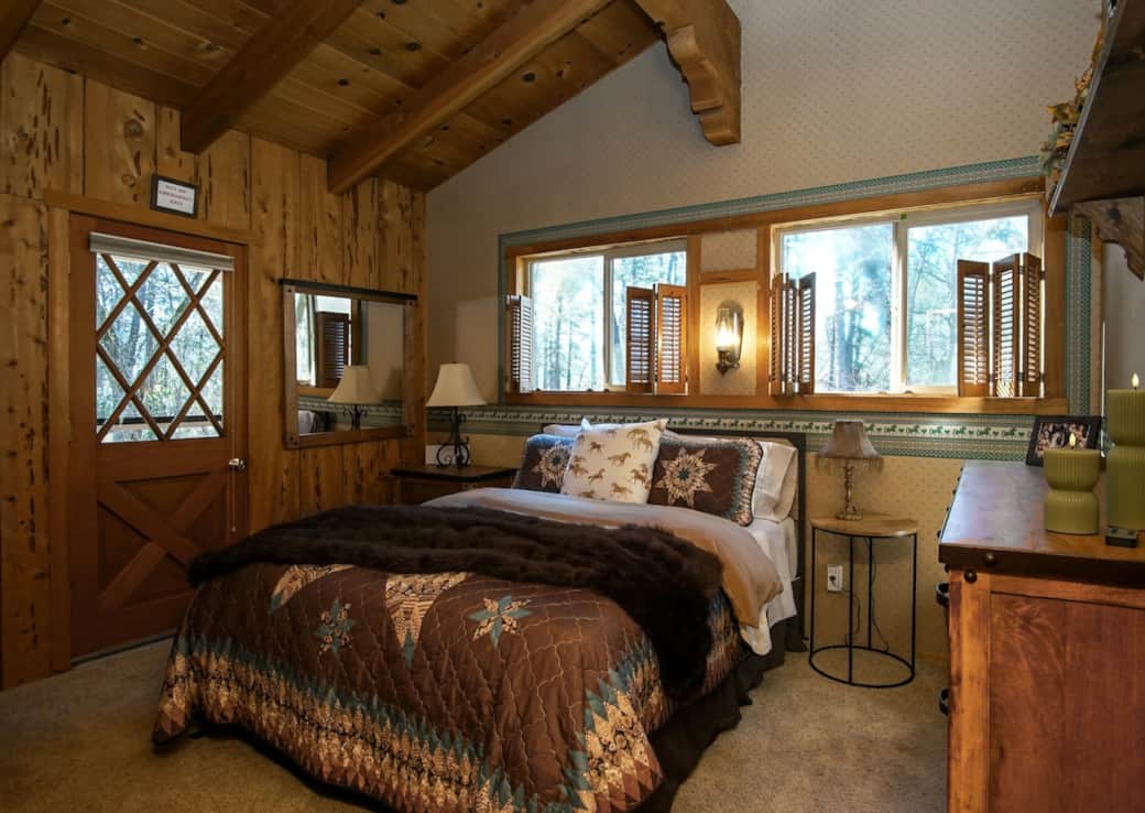 Unwind in The Bridle Room's rustic elegance. This cozy, wood-paneled sanctuary features a plush queen bed under vaulted exposed beams, classic Western decor, and custom window shutters. With a private exterior door, it’s the ultimate retreat.