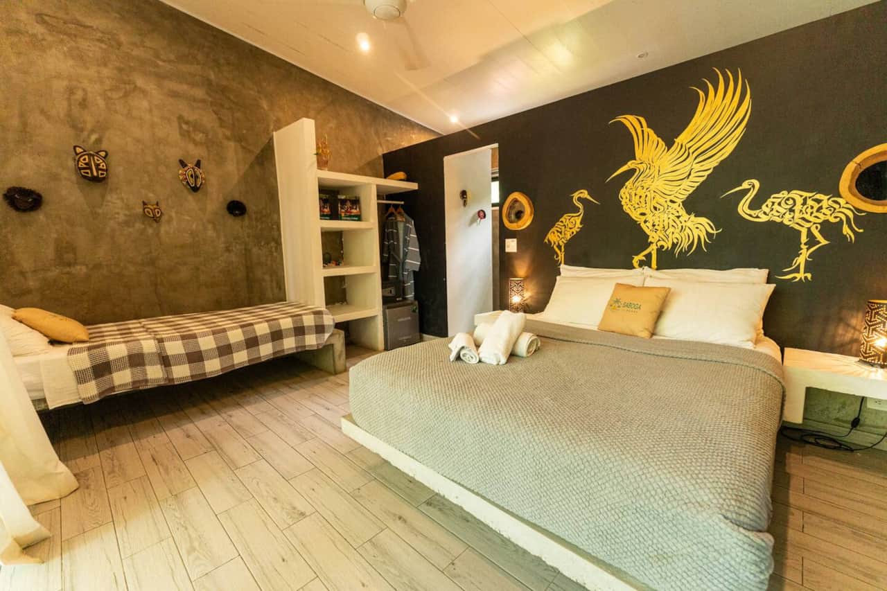 Máscaras en pared de concreto, mural dorado de aves y dos camas en la Deluxe Suite, Saboga Lodge