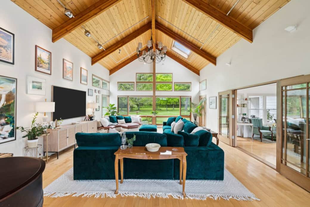 This light-filled great room is the heart of the home—featuring soaring cathedral ceilings, a wall of windows framing lush green views, and deep velvet and leather sofas perfect for lounging, reading, or movie nights.