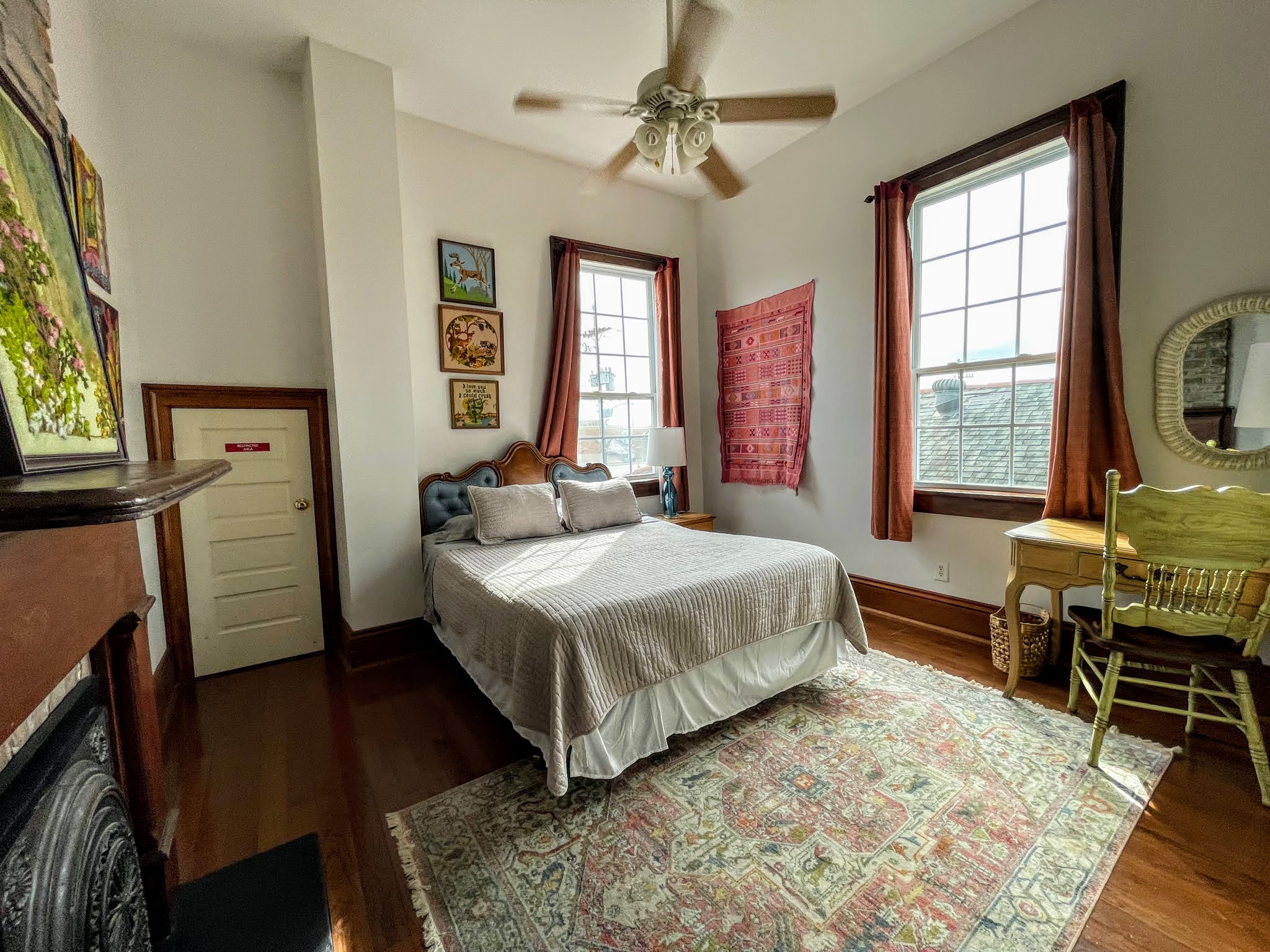 A wide view of a guest room with a bed, desk, rug, fireplace, and ceiling fan.