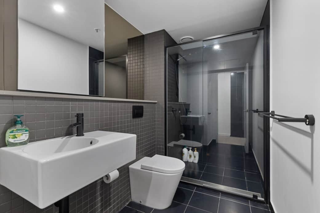 The bathroom features a sleek sink, a wall-mounted toilet, and a glass-enclosed shower. The dark tiles contrast with the white fixtures, creating a sophisticated look. A well-lit space that combines functionality with contemporary style.