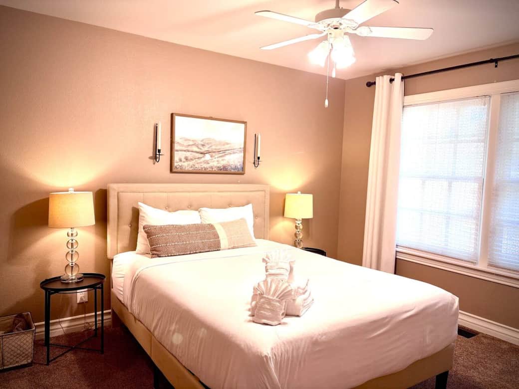 Main Floor Bedroom 2 (Queen Bed) – Warm, traditional mountain decor with plenty of closet space. Soothing beige decor with bedside lamps, ceiling fan, and large windows offering natural light. Perfect blend of comfort & style for your relaxing stay.