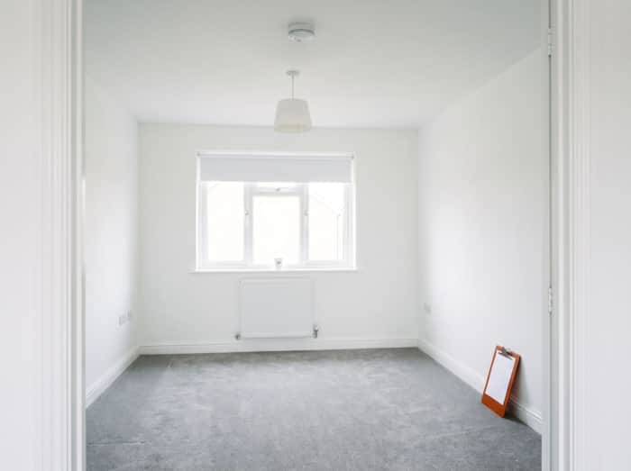 An empty, freshly painted room with grey carpet, a white blind and a smoke alarm