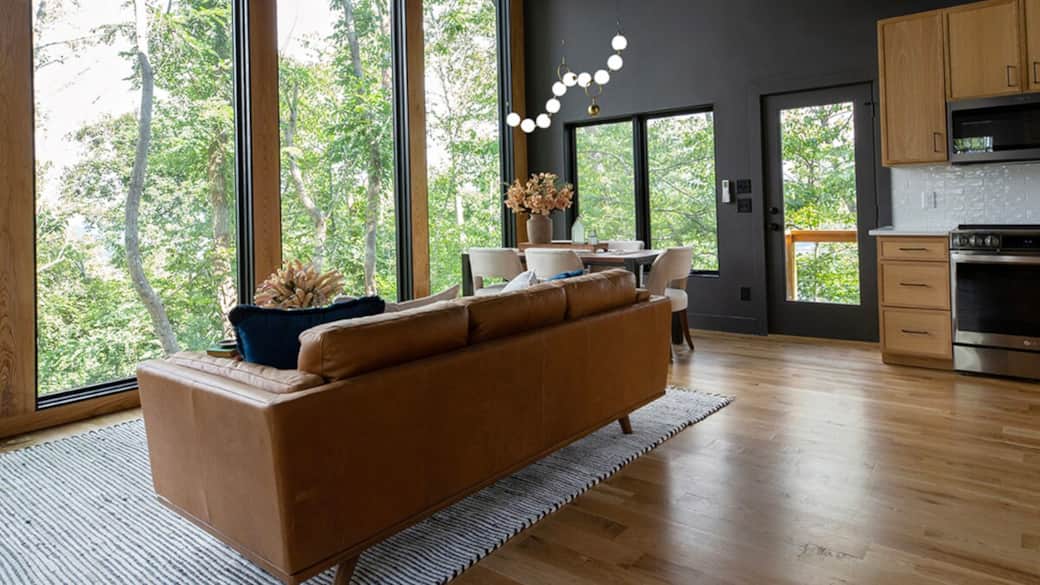 Bright and inviting open-living space featuring a living room, dining area and partial view of the kitchen. A full window wall frames the woods outside, bringing in natural light, creating a peaceful backdrop for your stay. Bright and inviting open-living space featuring a living room, dining area and partial view of the kitchen. A full window wall frames the woods outside, bringing in natural light, creating a peaceful backdrop for your stay.