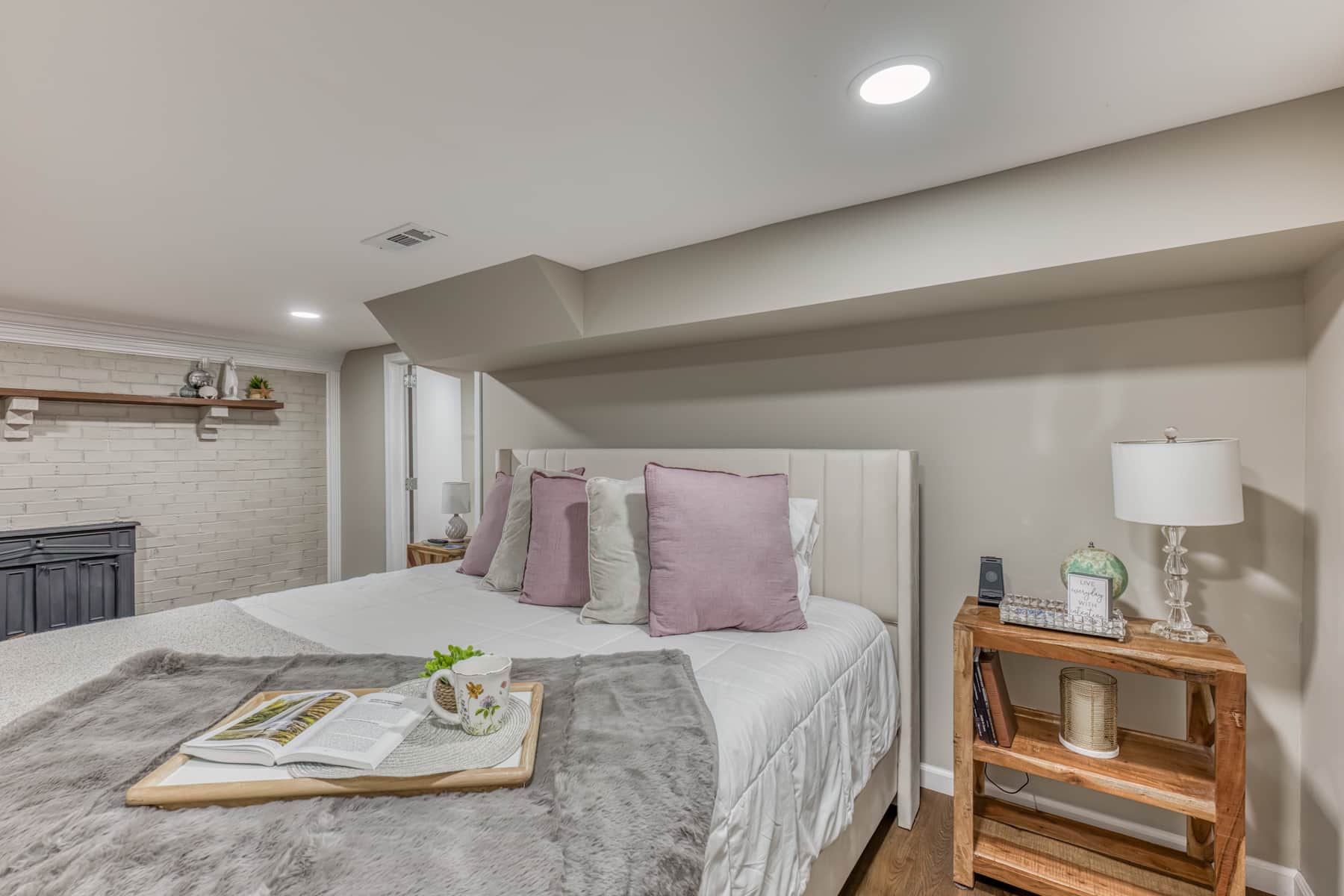 Main-level king primary suite with smart TV, private bathroom, decorative brick fireplace feature wall, spacious layout, and luxury bedding.