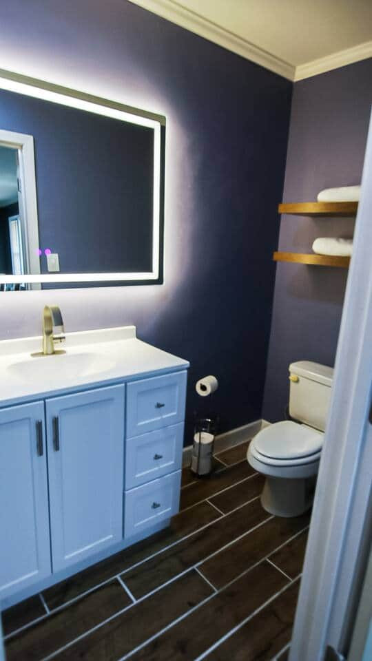 (Bathroom 2) A crisp white vanity with faucet sits beneath a sleek backlit mirror. Wood tile floors, floating timber shelves, and deep navy walls add warmth and character—turning this bath into a spa-like retreat. (Bathroom 2) A crisp white vanity with faucet sits beneath a sleek backlit mirror. Wood tile floors, floating timber shelves, and deep navy walls add warmth and character—turning this bath into a spa-like retreat.