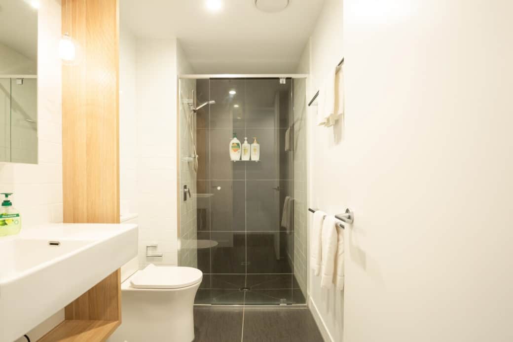 This bathroom is a model of cleanliness and efficiency, equipped with a modern sink, toilet, and shower. The white fixtures and tiles create a fresh and crisp look, while the well-lit space ensures a pleasant experience for guests.