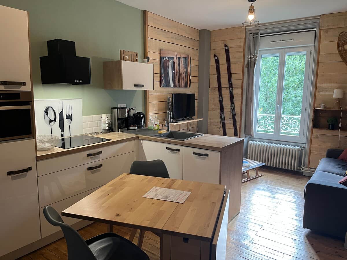 Cuisine moderne avec meubles blancs et bois, équipée d’appareils électroménagers. Table en bois clair avec chaises grises. Décor mural en bois, skis vintage, fenêtre lumineuse donnant sur des arbres.