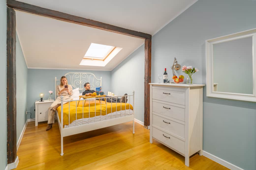 Sypialnia z białym, metalowym łóżkiem pod oknem dachowym. 