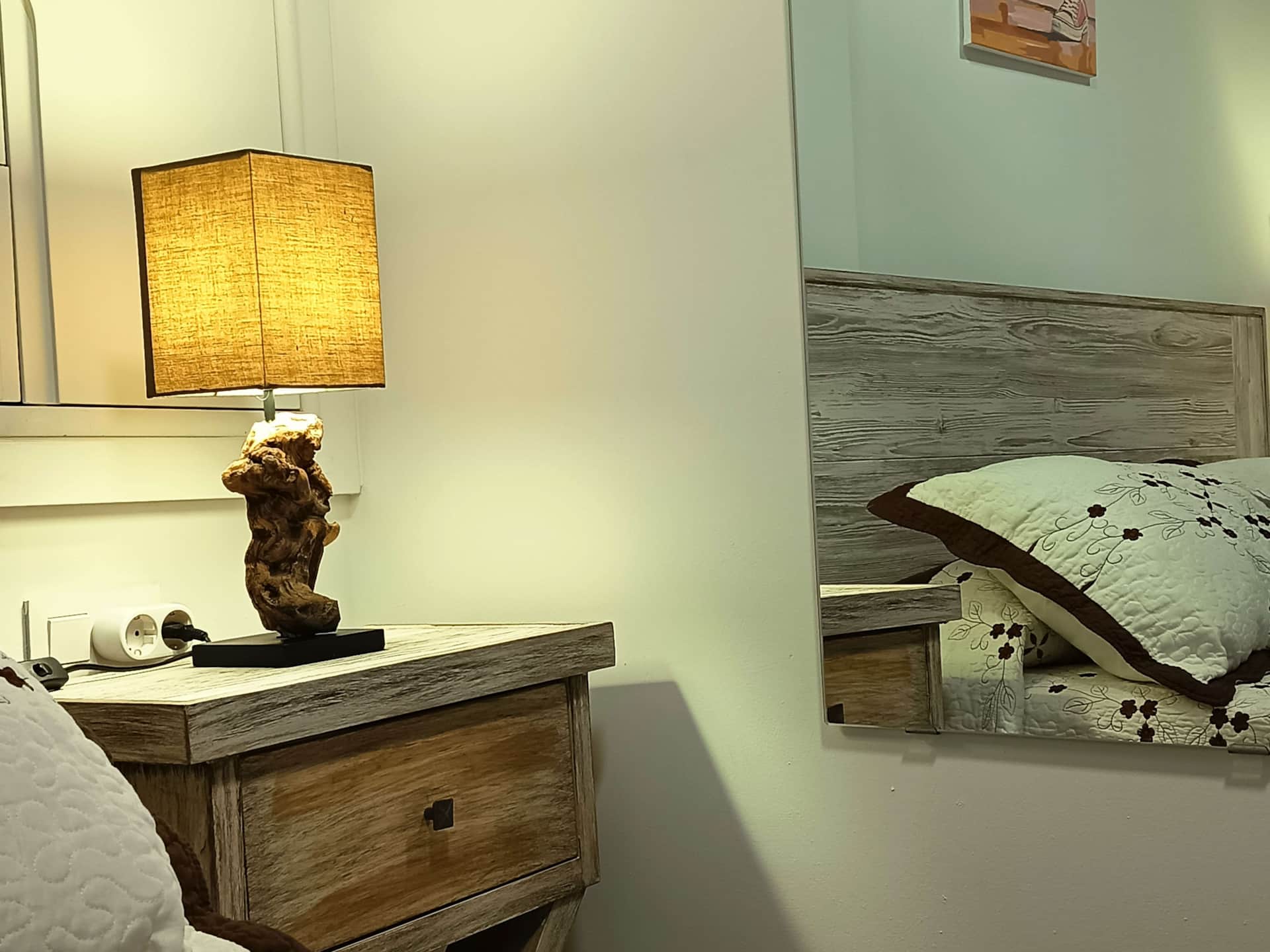 Mesilla de noche de madera con lámpara de base rústica y pantalla rectangular, junto a cama con cabecero de madera y colcha floral