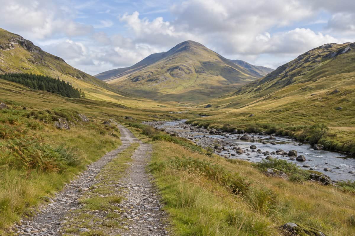 AI illustration: track and stony river winding into a broad grassy glen