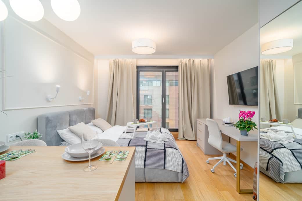 A spacious and bright studio room with a kitchenette, a large bed, and a desk by the window.
