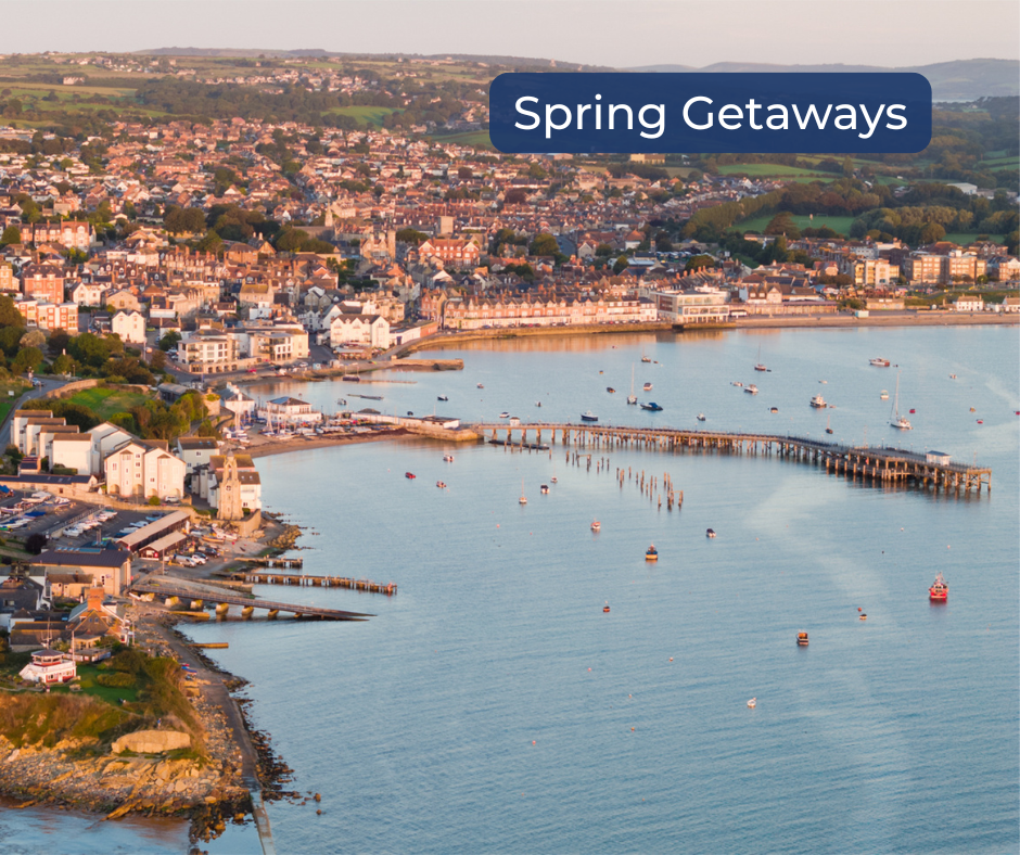 Drone photo of Swanage Bay in spring with clear blue water, sandy shoreline, and the text ‘Spring Getaways’ in the corner